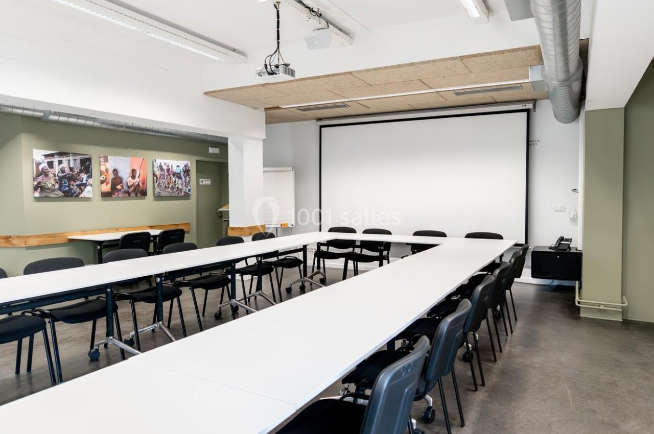 Salle de réunion lumineuse avec tables disposées en U, chaises noires, écran de projection et décorations murales.