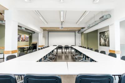 Personnes assises à des tables sur une terrasse en bois, entourée de verdure et d'immeubles modernes en arrière-plan.