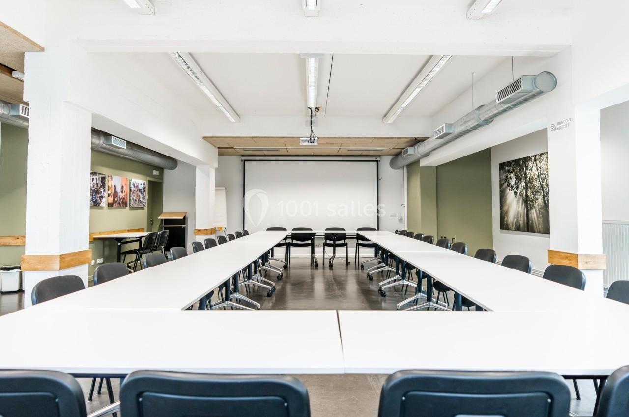 Salle de réunion lumineuse avec des tables disposées en U, des chaises noires et un écran de projection au fond.