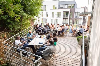 Personnes assises à des tables sur une terrasse en bois, entourée de verdure et d'immeubles modernes en arrière-plan.