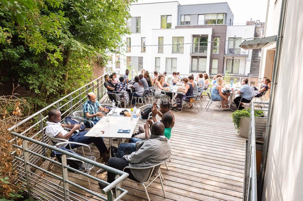 Personnes assises à des tables sur une terrasse en bois, entourée de verdure et d'immeubles modernes en arrière-plan.