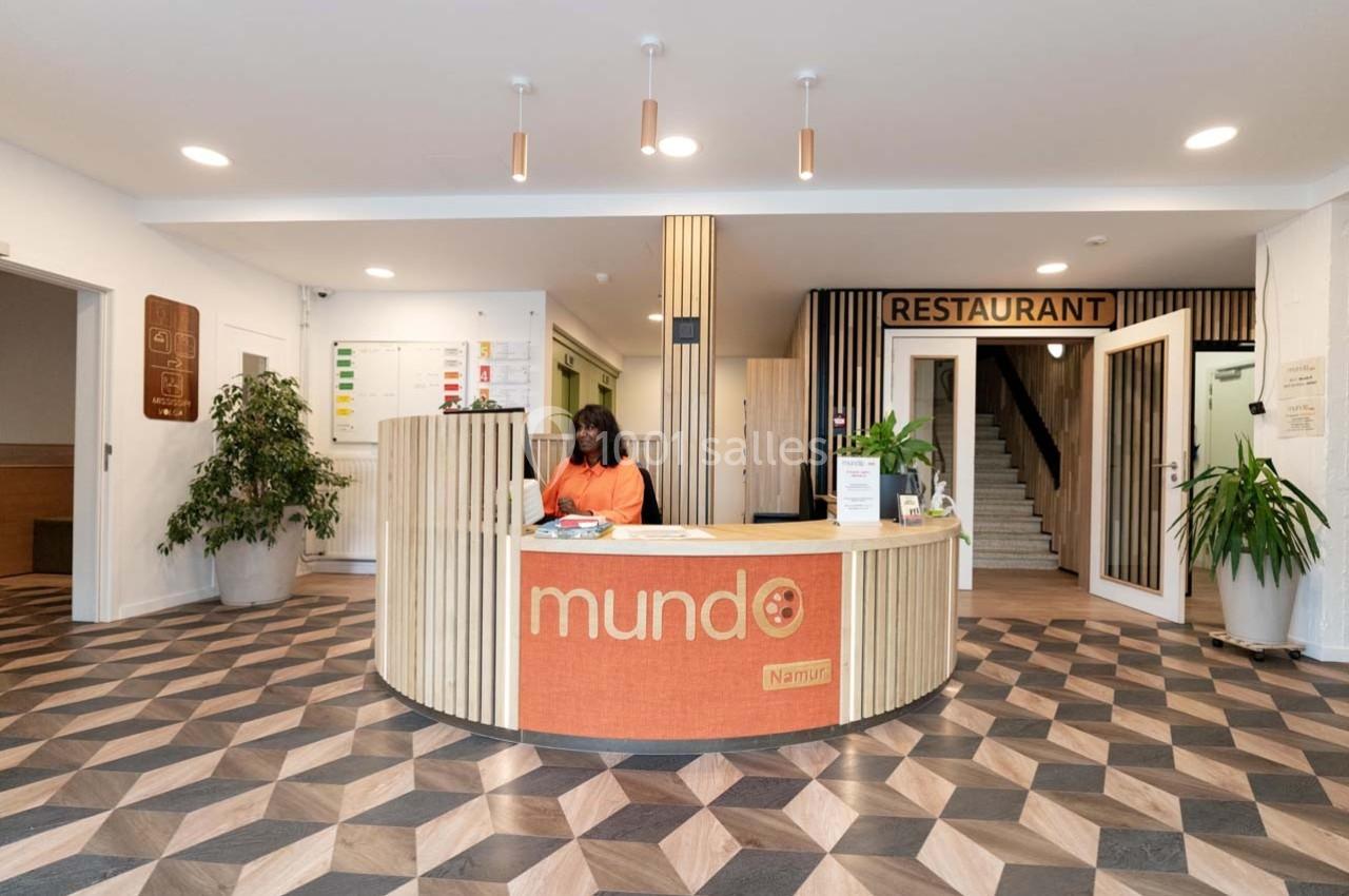 Accueil avec comptoir en bois, employée assise, plantes décoratives et signalétique vers un restaurant en arrière-plan.