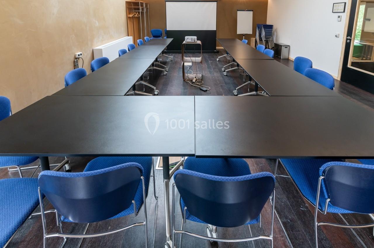 Salle de réunion avec tables noires disposées en U, chaises bleues, projecteur et écran de projection à l'arrière.
