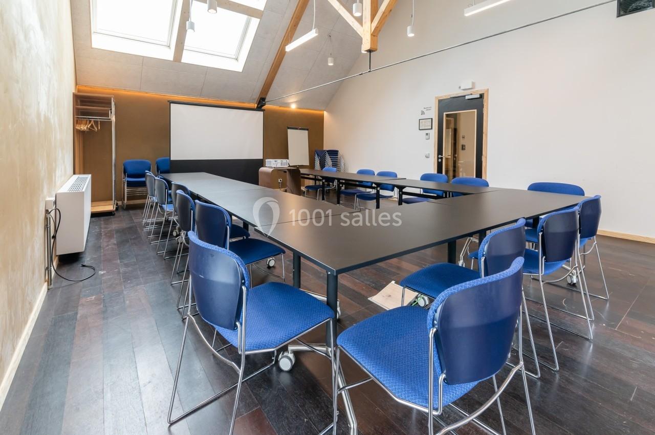 Salle de réunion lumineuse avec tables disposées en U, chaises bleues et écran de projection au fond.