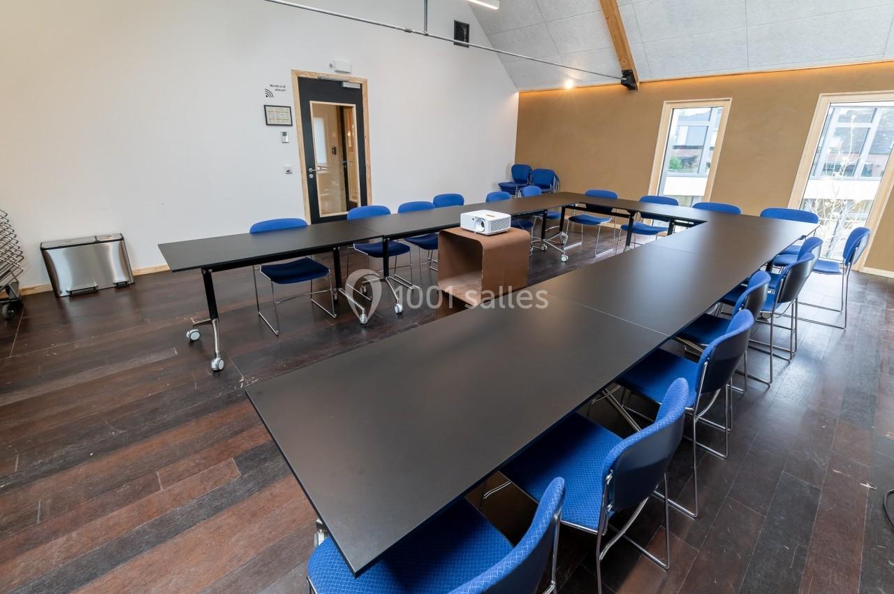 Salle de réunion lumineuse avec tables noires en U, chaises bleues et projecteur sur un meuble central.