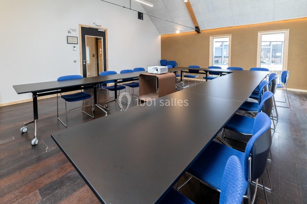 Salle de réunion avec tables noires disposées en U, chaises bleues et projecteur au centre, éclairée par des fenêtres.