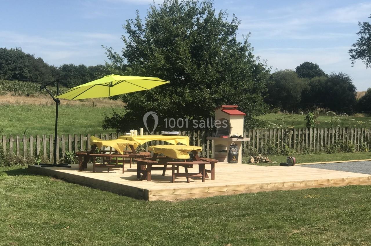 Terrasse en bois avec tables, parasol vert et barbecue, entourée de pelouse et d'arbres sous un ciel dégagé.