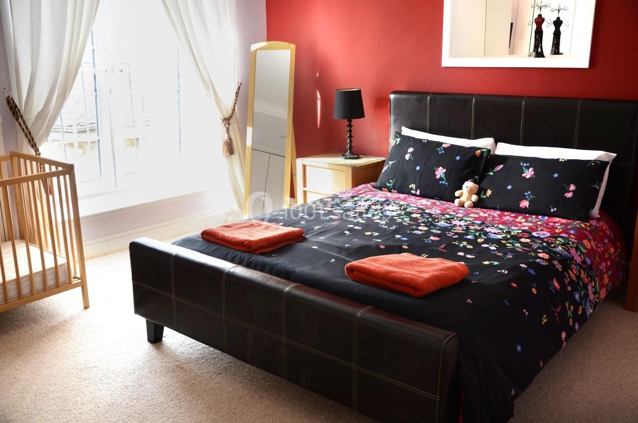 Chambre avec lit double, draps à motifs floraux, lit bébé en bois, murs rouges et fenêtre avec rideaux blancs.