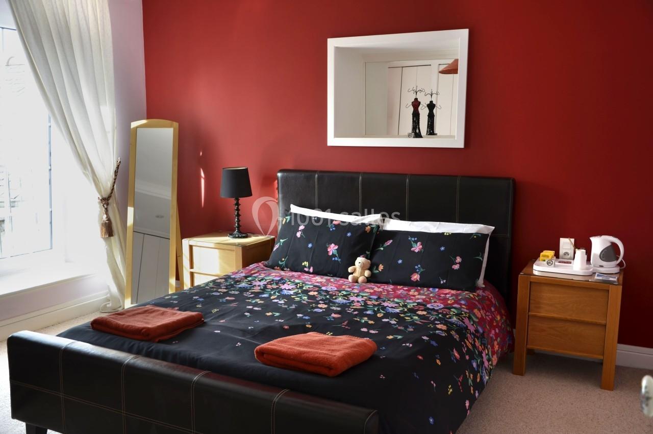 Chambre avec un lit double, draps à motifs floraux, murs rouges, miroir, table de chevet et lumière naturelle.