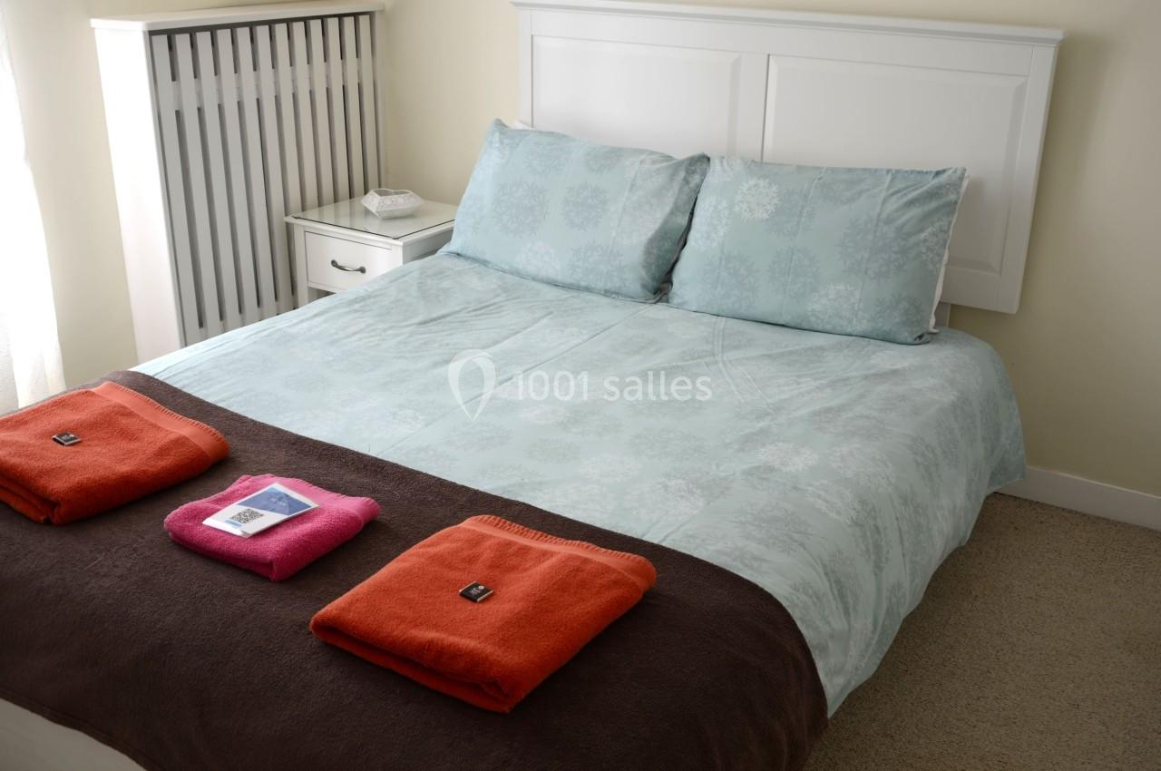 Lit double avec draps bleus, deux oreillers, couverture marron et serviettes pliées orange et rose sur le dessus.