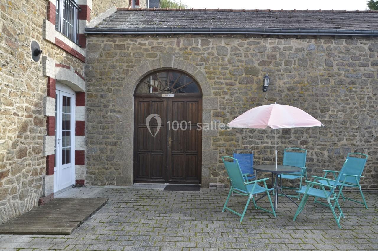 Cour pavée avec une table ronde, quatre chaises turquoise, un parasol rose et une façade en pierre avec une porte en bois.
