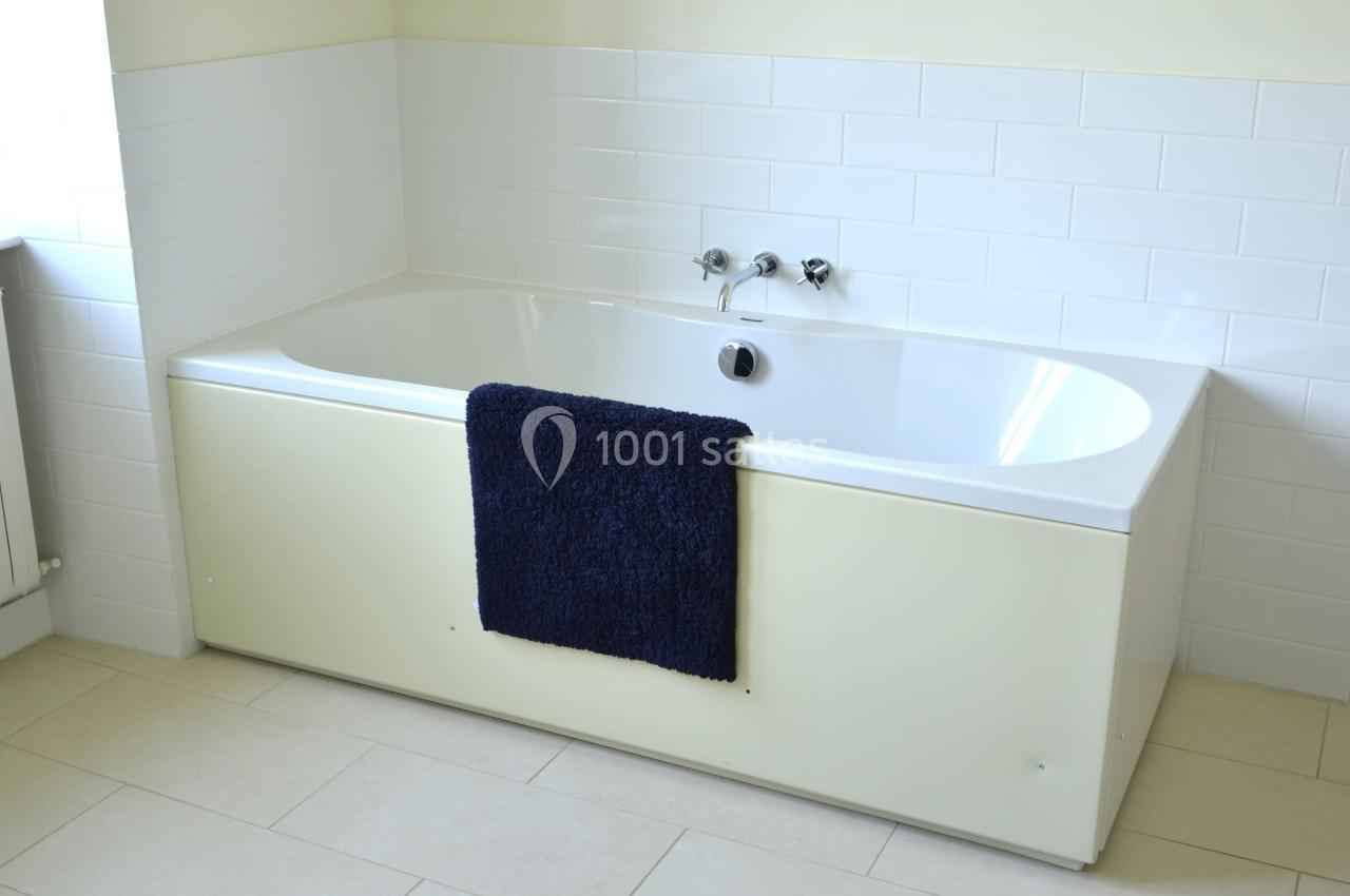 Baignoire blanche avec un tapis bleu foncé suspendu, entourée de carrelage blanc et d'un sol beige.