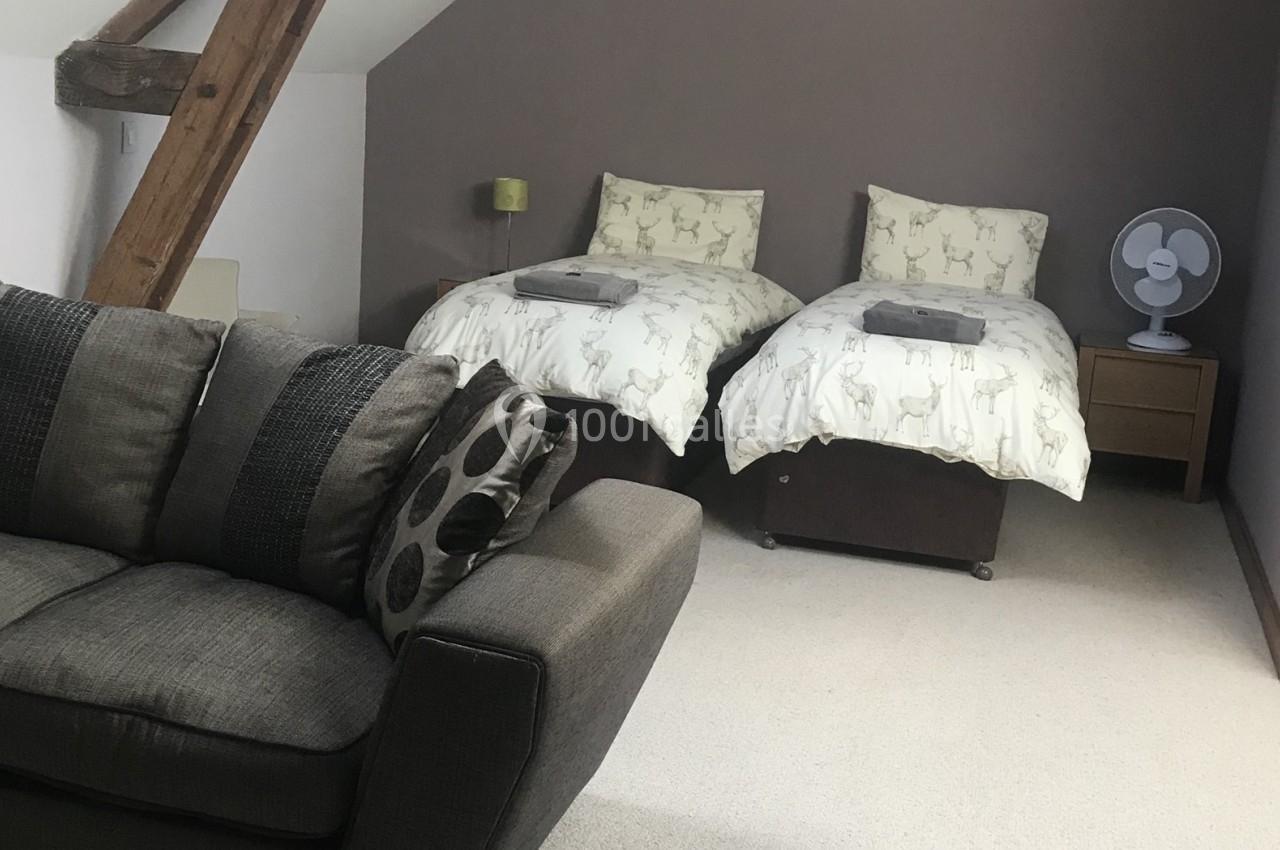 Chambre avec deux lits simples, draps à motifs de cerfs, canapé gris au premier plan et ventilateur sur une table de chevet.