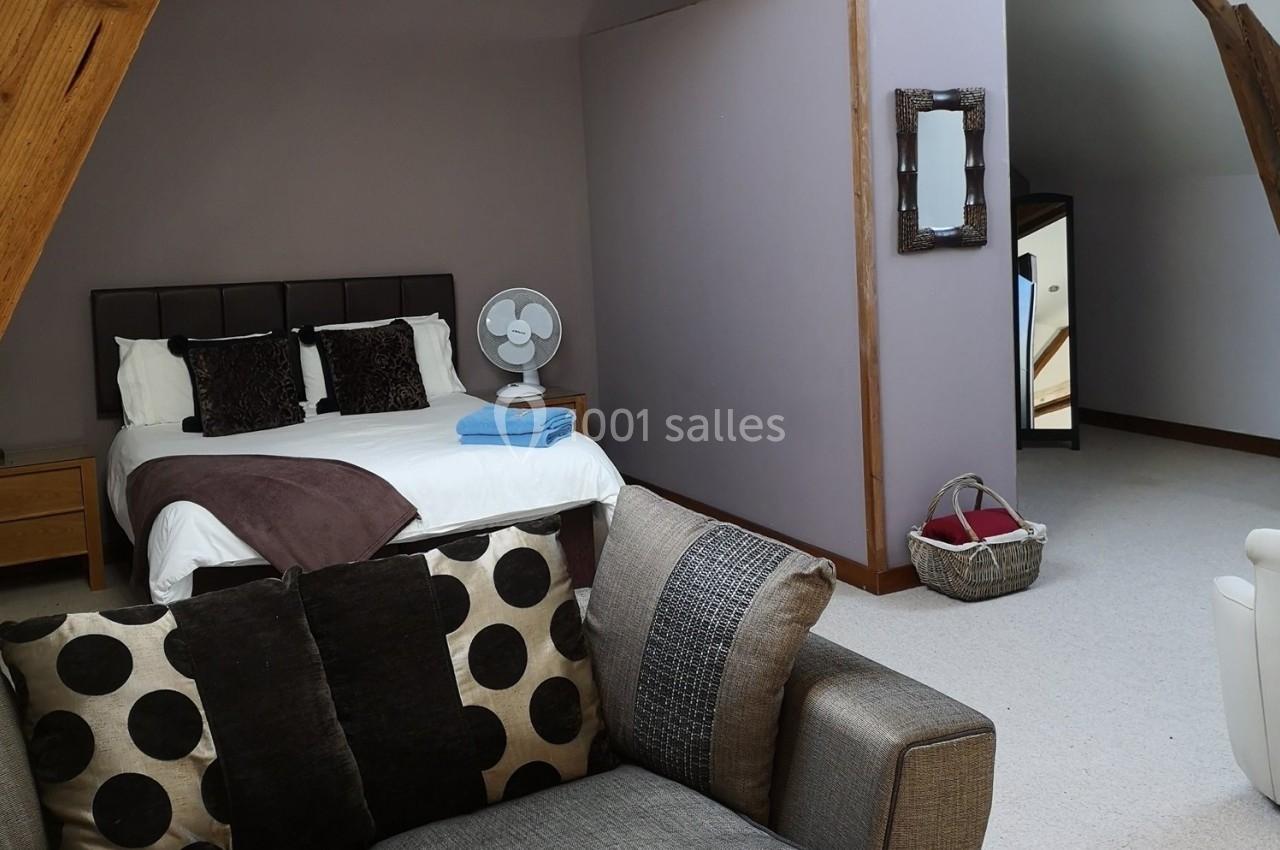 Chambre mansardée avec un lit double, un ventilateur sur une table de chevet, un canapé et un panier au sol.