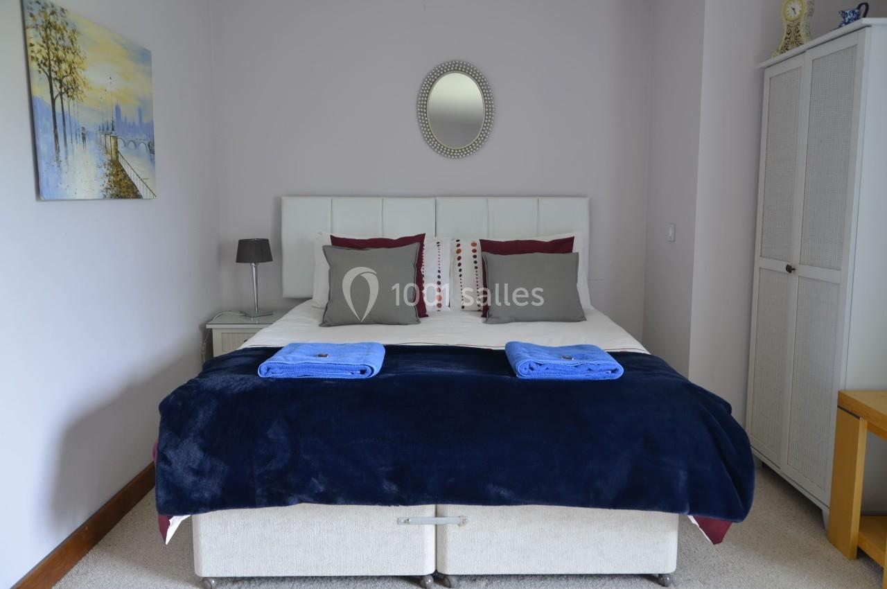 Lit double avec tête de lit blanche, couvertures bleues pliées, coussins décoratifs, dans une chambre lumineuse.