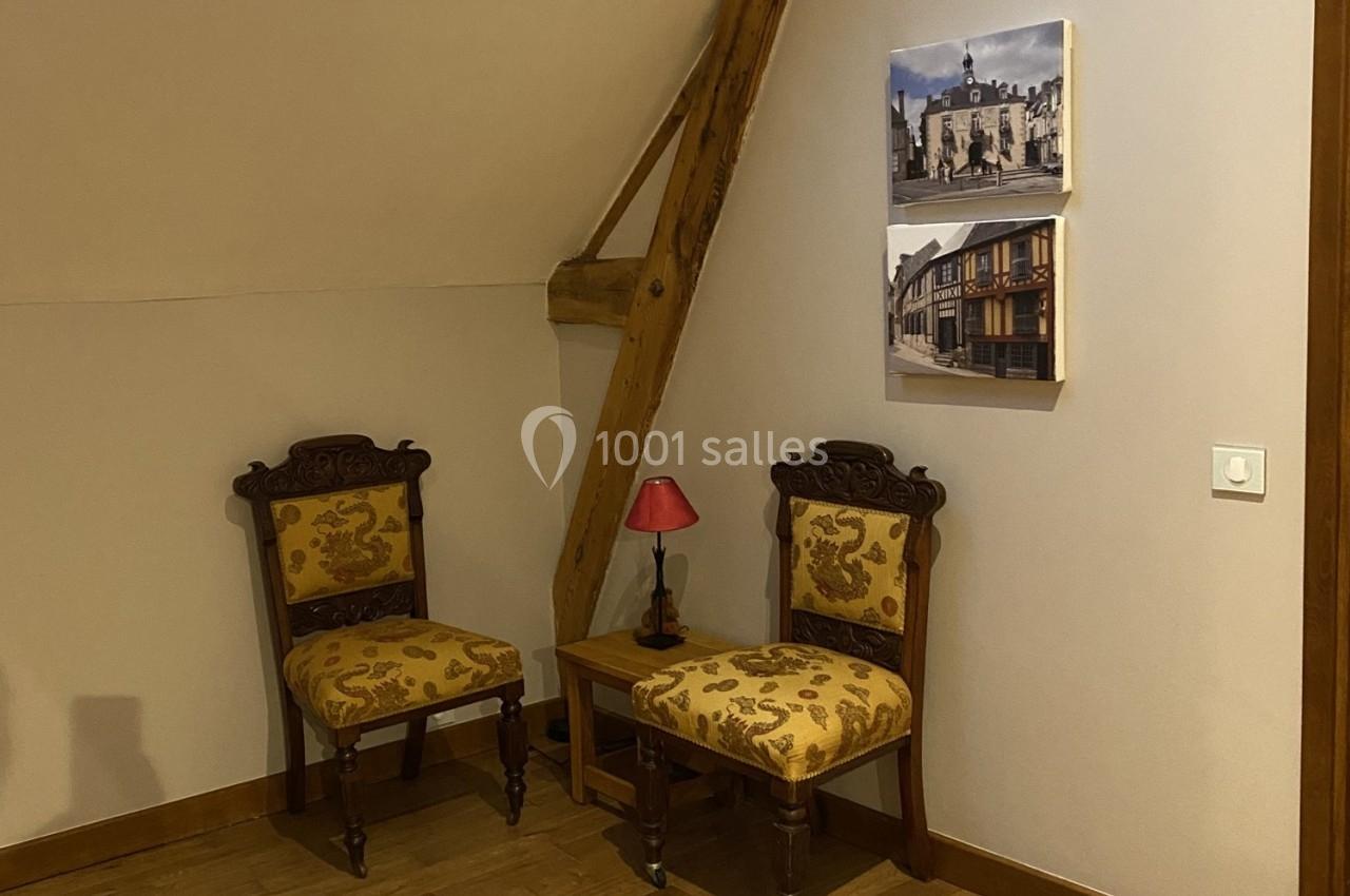 Deux chaises anciennes avec motifs floraux, une petite table avec lampe rouge, et deux cadres accrochés au mur.
