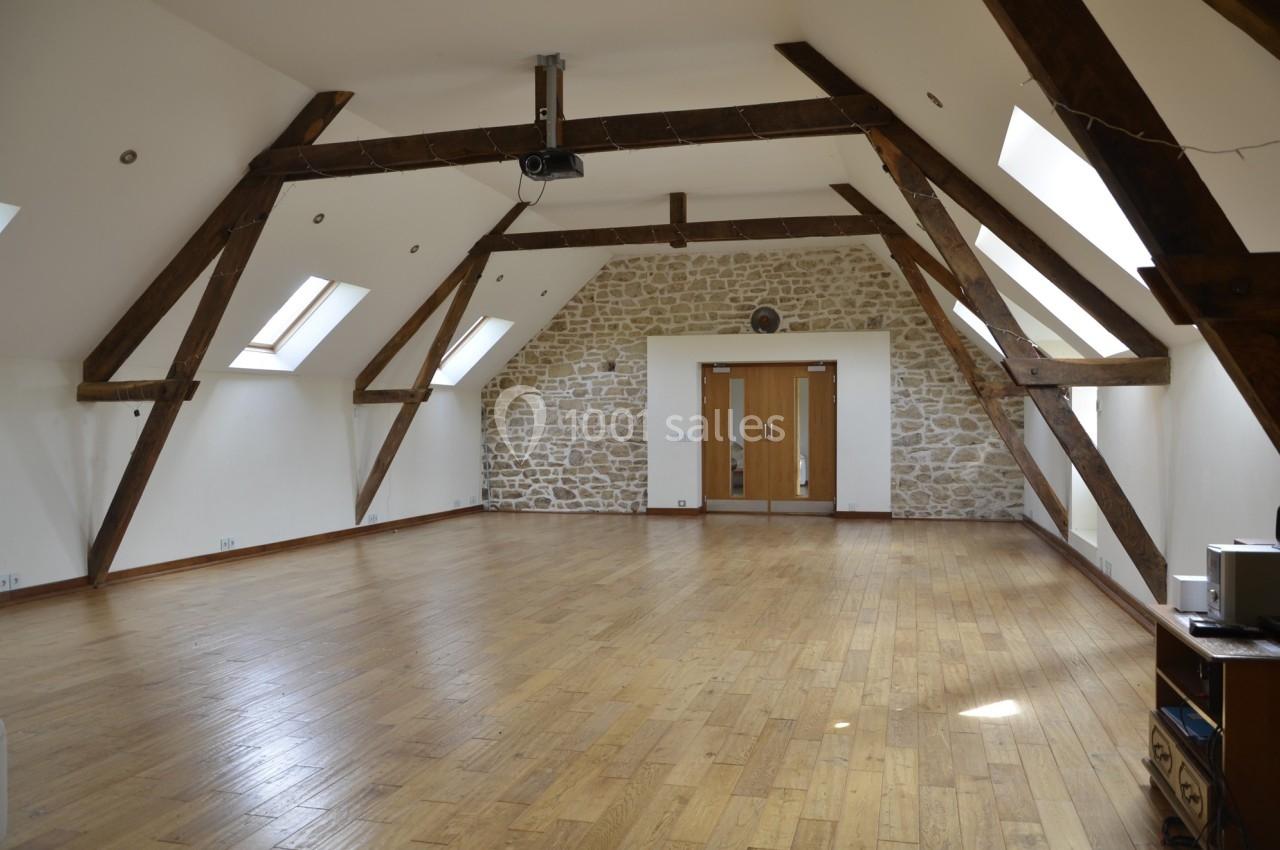 Salle spacieuse avec poutres apparentes, parquet en bois, murs en pierre et fenêtres de toit laissant entrer la lumière.