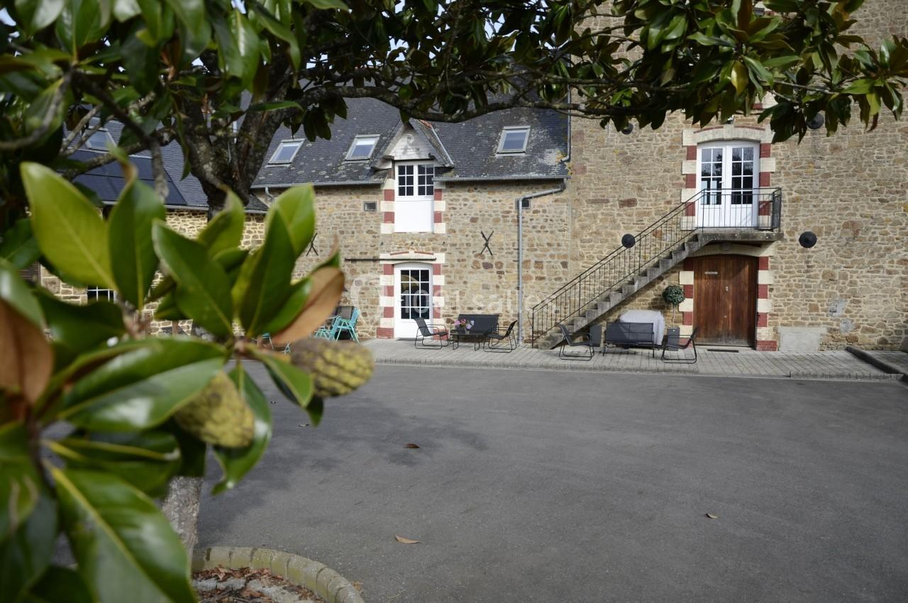 Façade en pierre d'une maison avec escalier extérieur, vue partiellement encadrée par des feuilles d'arbre au premier plan.