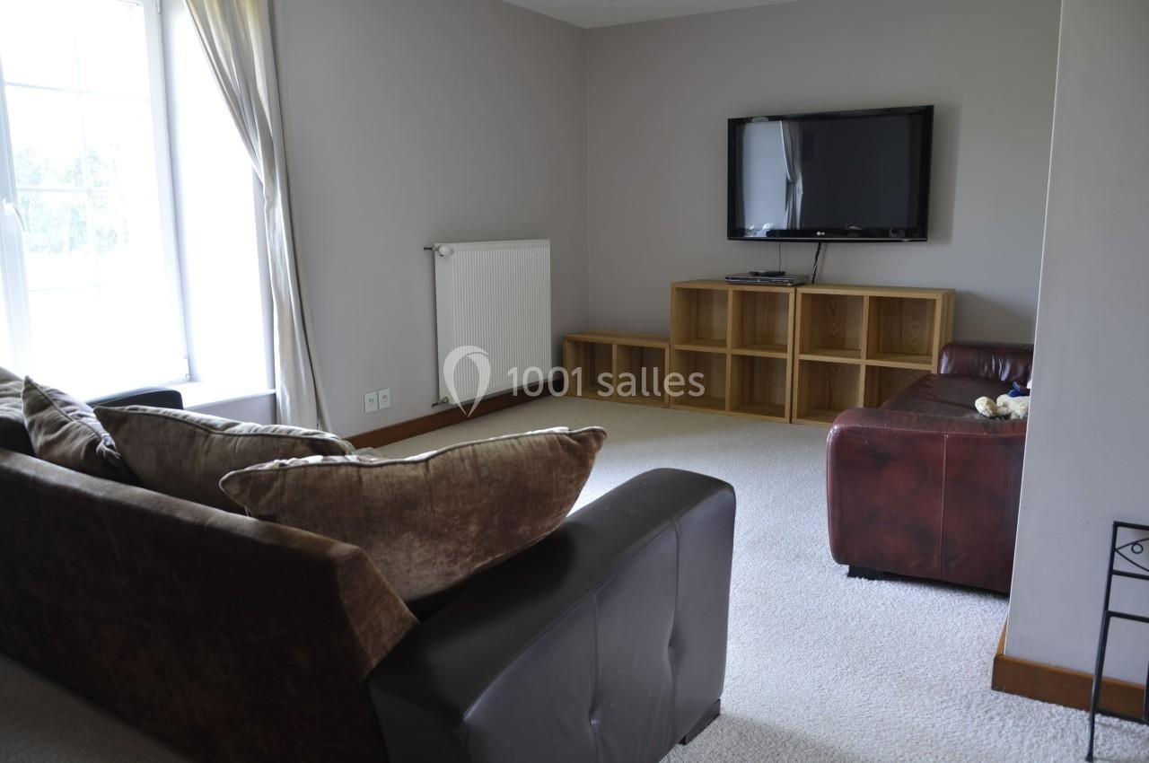 Salon avec canapé, fauteuil en cuir, étagère en bois, télévision murale et grande fenêtre laissant entrer la lumière.