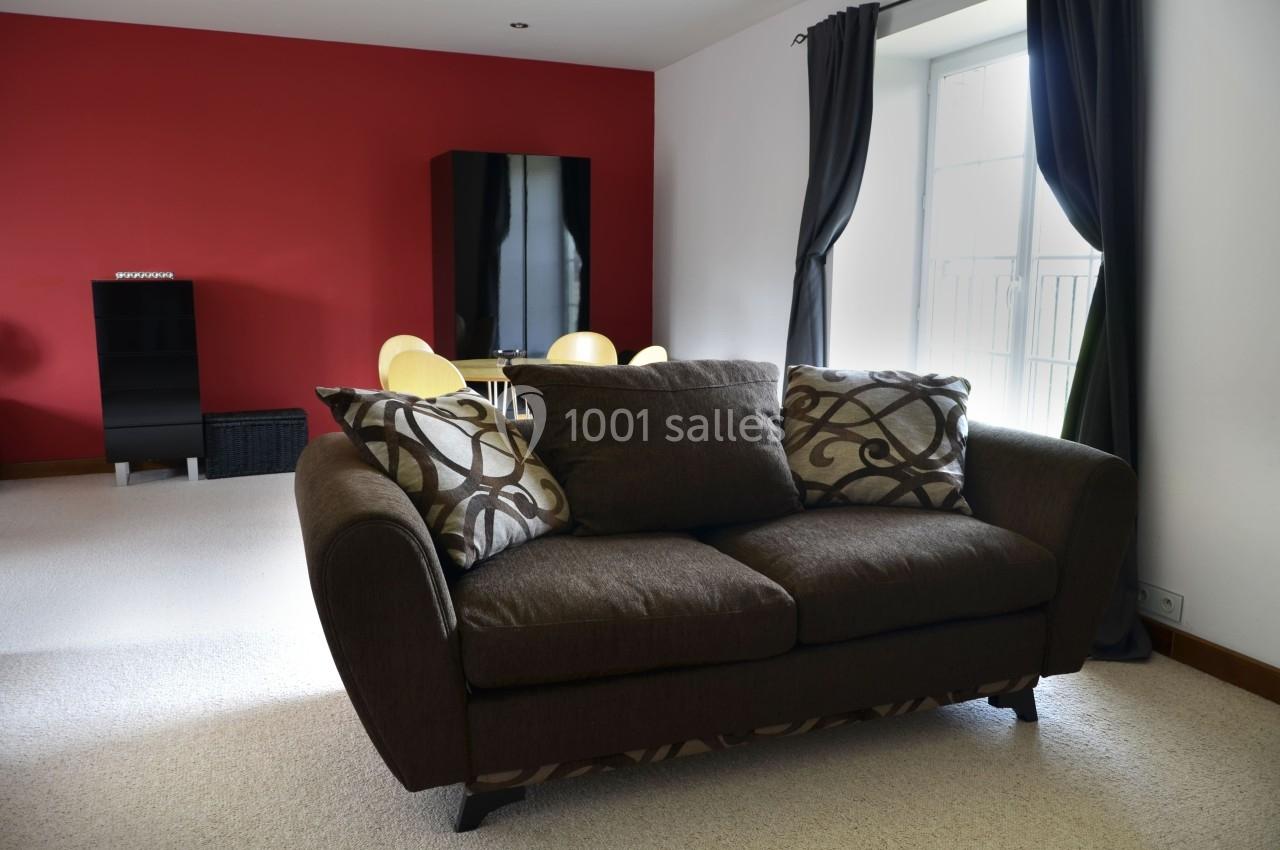 Canapé marron avec coussins décoratifs dans un salon lumineux, murs rouges et blancs, table et chaises en arrière-plan.