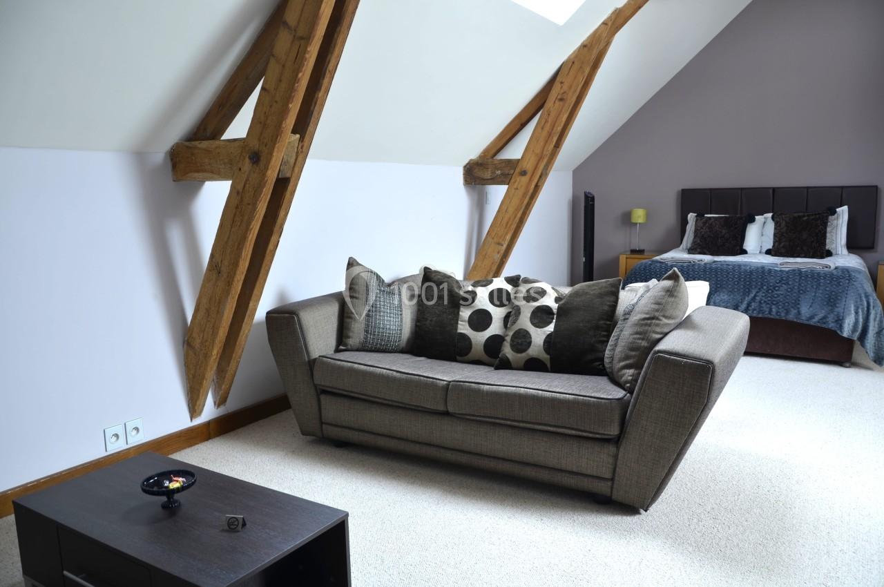 Canapé gris avec coussins décoratifs dans une chambre mansardée, lit double en arrière-plan.