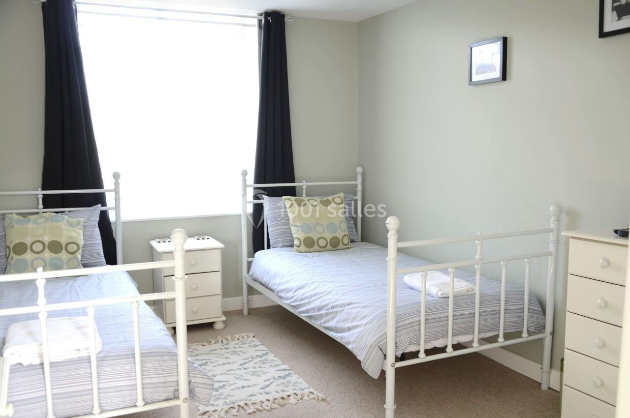 Deux lits simples avec linge rayé, table de chevet entre eux, fenêtre avec rideaux noirs et mur clair décoré d'un cadre.