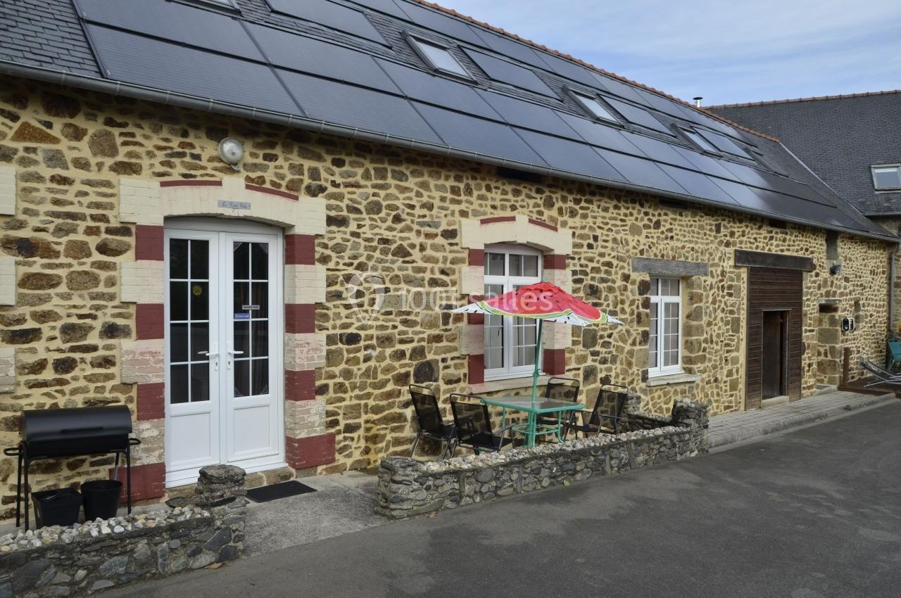 Façade en pierre d'une maison avec panneaux solaires, porte blanche, table extérieure et parasol rouge.