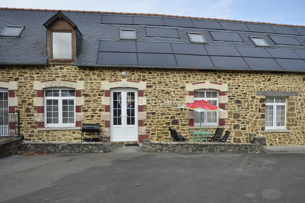 Façade en pierre d'une maison avec fenêtres blanches, toit en ardoise et panneaux solaires, table et parasol devant.