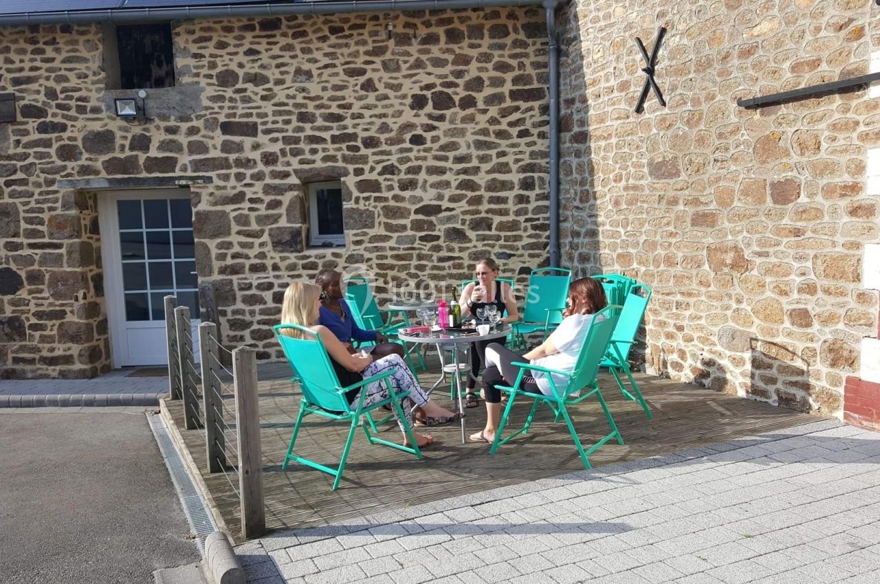 Quatre personnes assises autour d'une table sur une terrasse en bois, devant une maison en pierre.