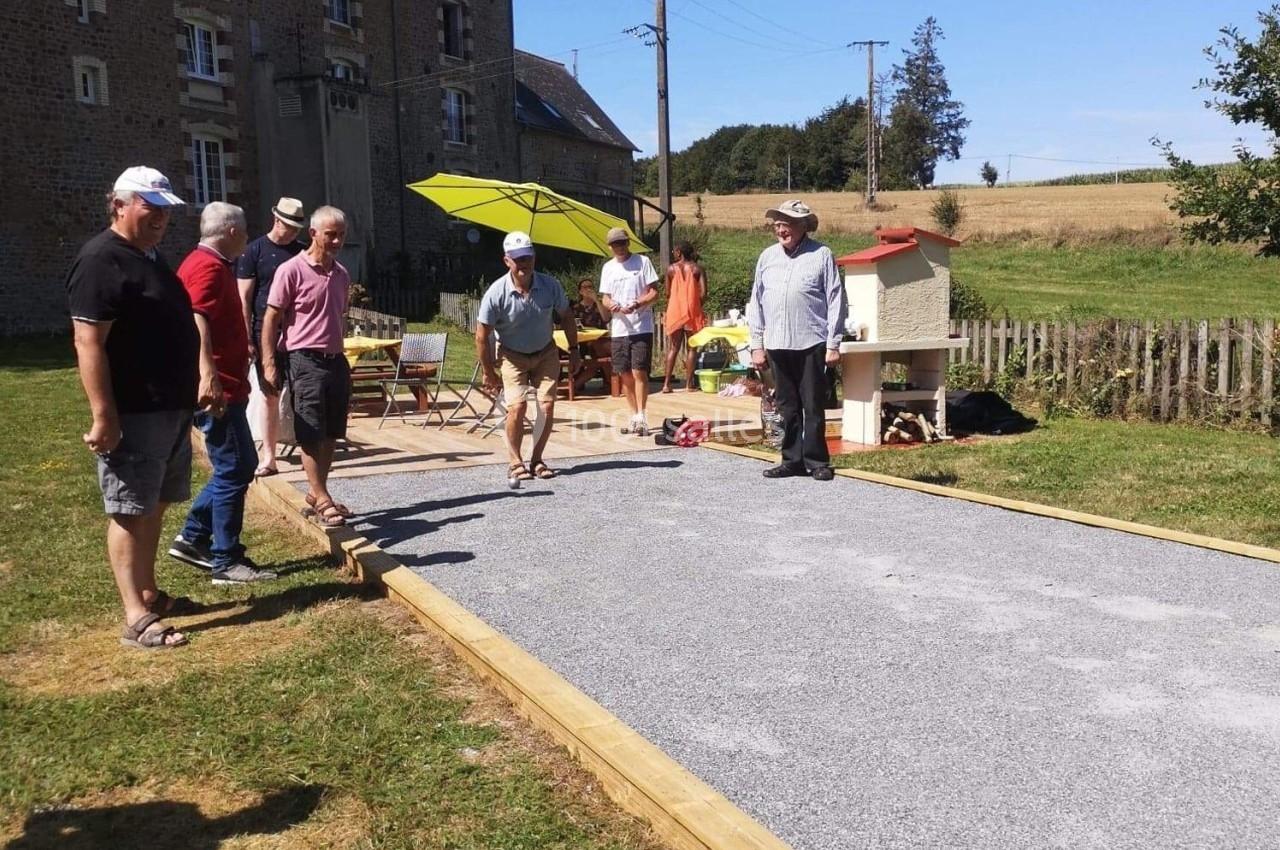 Des personnes jouent à la pétanque sur un terrain aménagé en plein air, près d'une maison en pierre et d'un jardin.