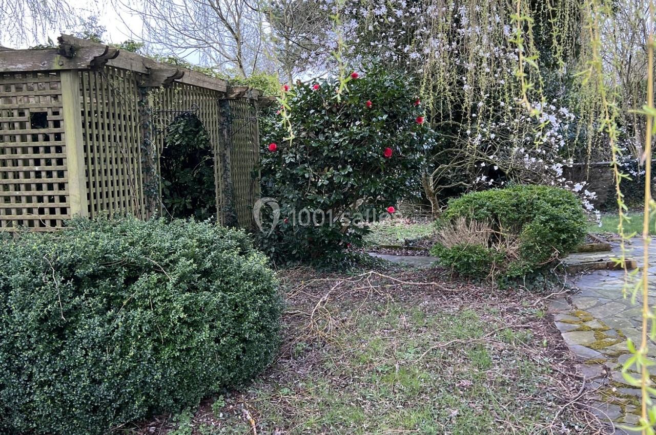 Pergola en bois entourée de buissons, d'arbustes fleuris et d'un chemin pavé dans un jardin.