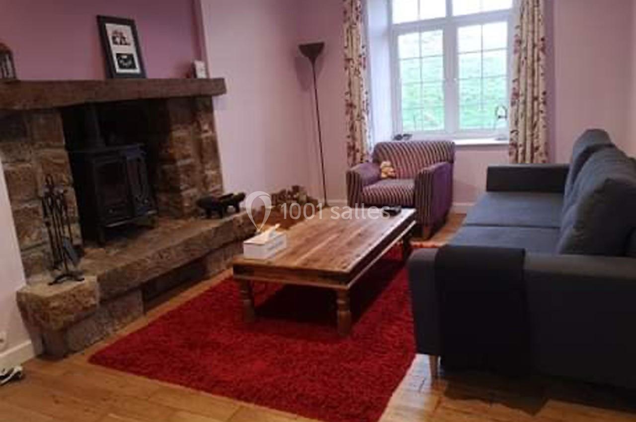 Salon avec murs roses, cheminée en pierre, poêle à bois, canapé gris, fauteuil rayé, table basse en bois et tapis rouge.