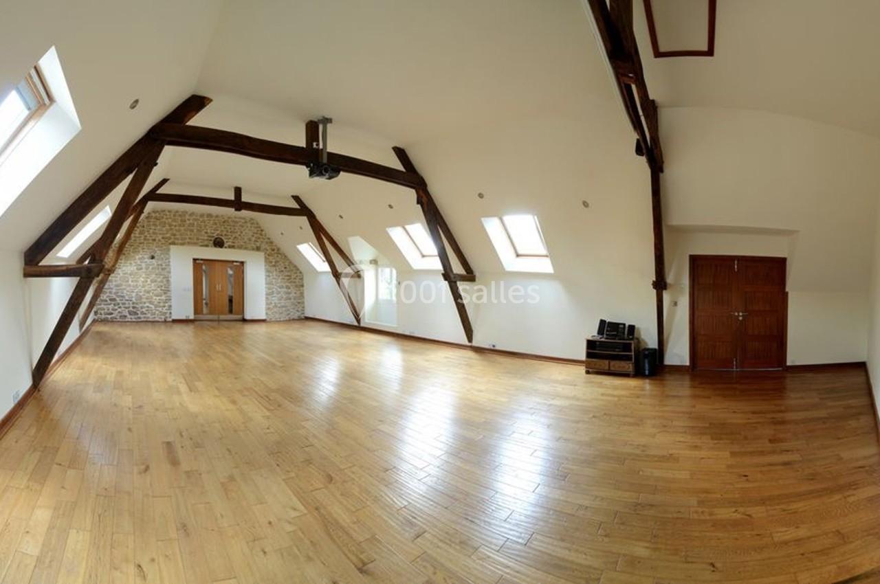 Grande pièce lumineuse avec parquet en bois, poutres apparentes, murs blancs et plusieurs fenêtres de toit.