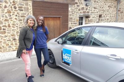 Deux femmes souriantes posent devant une voiture grise marquée ’France Bleu’, stationnée devant un bâtiment en pierre.