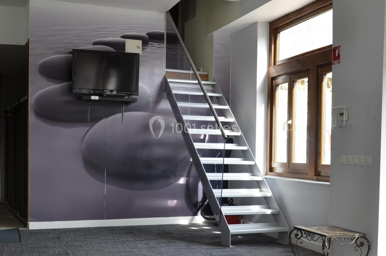 Escalier métallique intérieur près d'une fenêtre, avec un mur décoré de galets et une télévision fixée dessus.