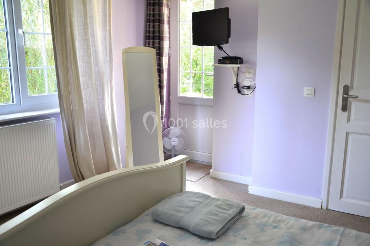 Chambre lumineuse avec lit, miroir, télévision murale, ventilateur au sol et fenêtres donnant sur un jardin.