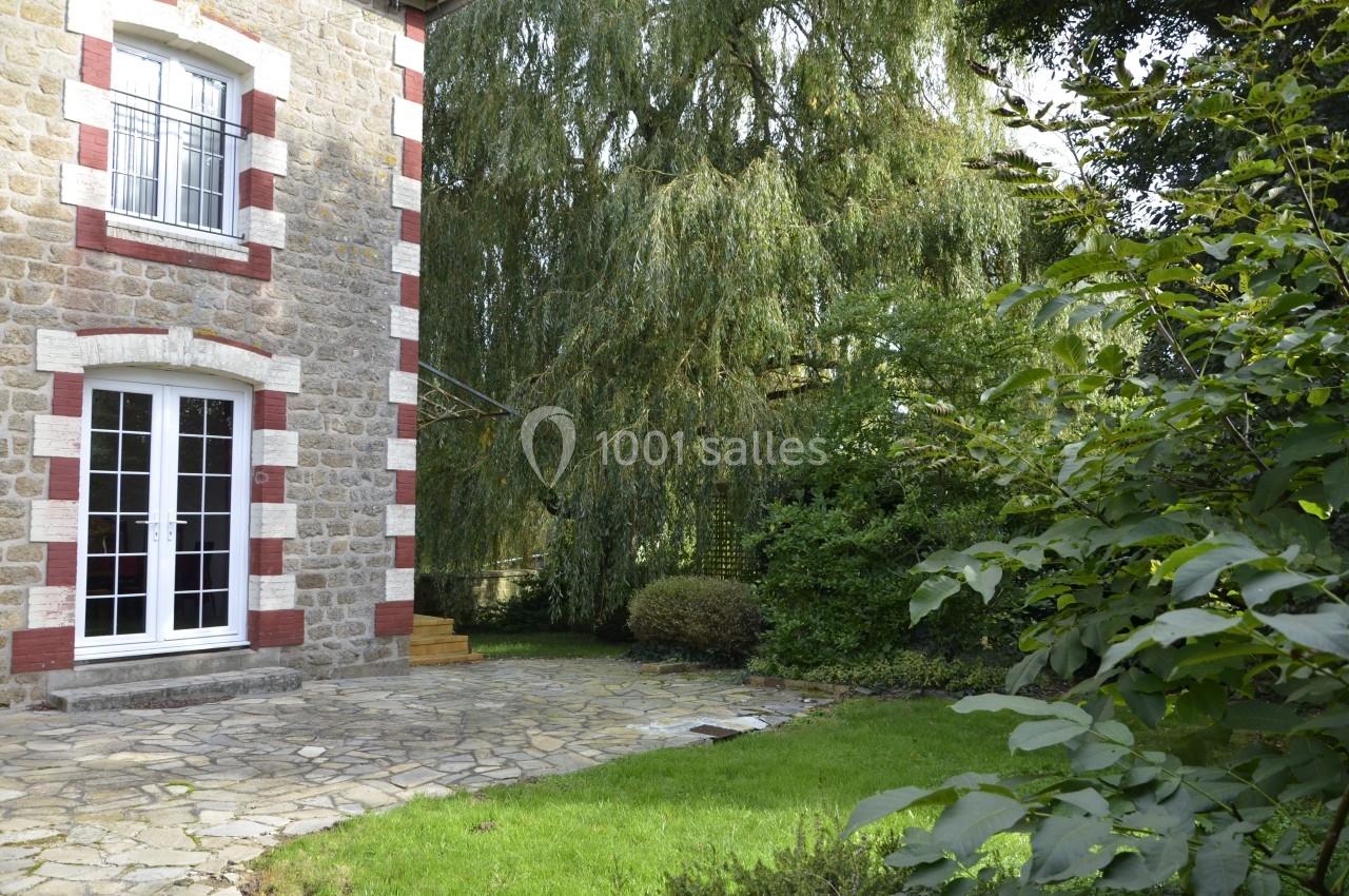 Façade en pierre d'une maison avec terrasse en pierre, jardin verdoyant et arbre imposant en arrière-plan.