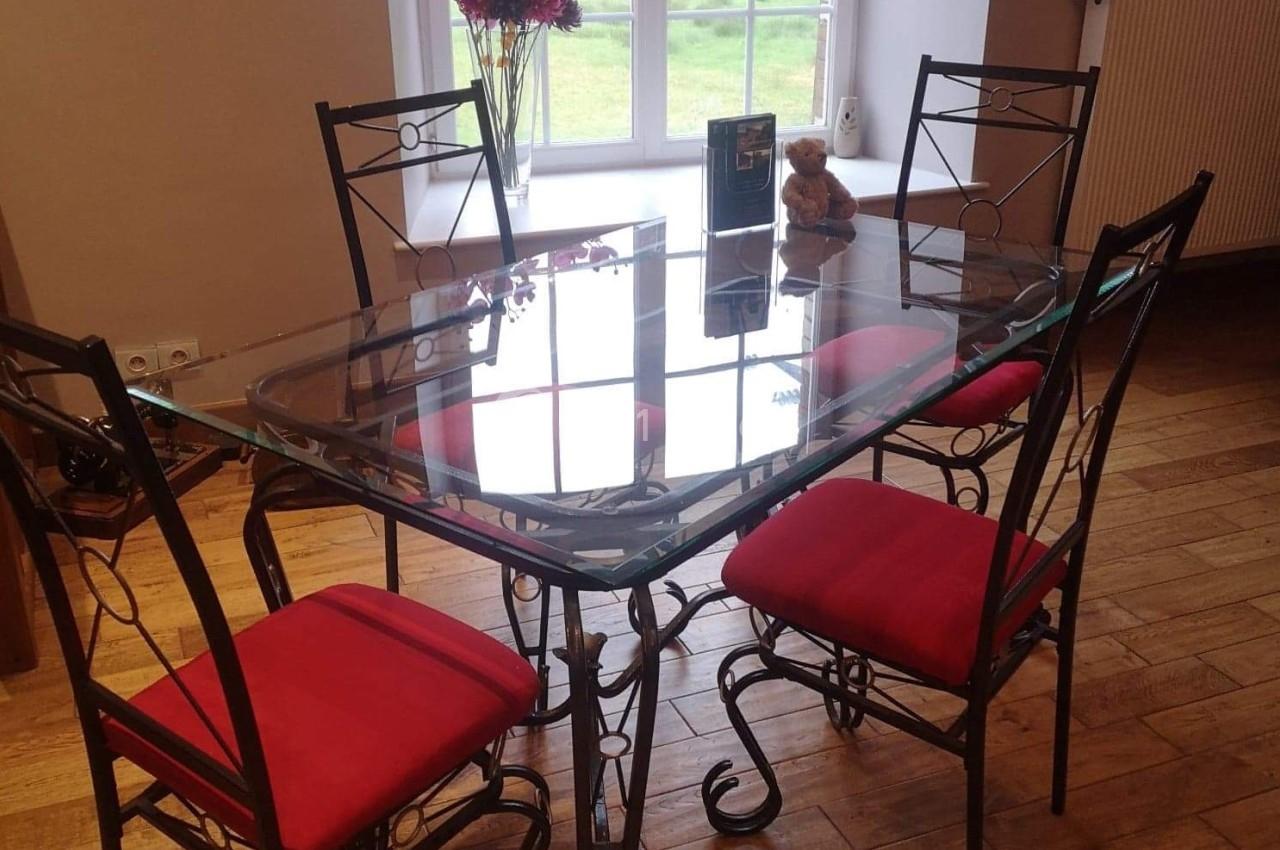 Table en verre avec structure en métal noir, entourée de quatre chaises à coussins rouges, dans une pièce lumineuse.