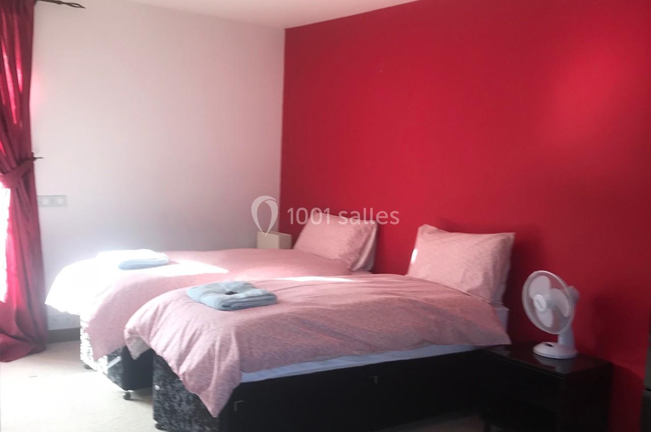 Deux lits simples avec draps roses dans une chambre lumineuse aux murs rouges et blancs, rideaux rouges et ventilateur sur…