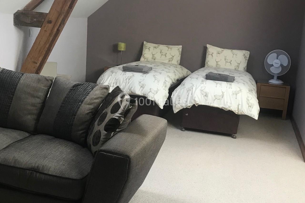 Chambre mansardée avec deux lits simples, canapé, table basse, ventilateur et poutres apparentes en bois.