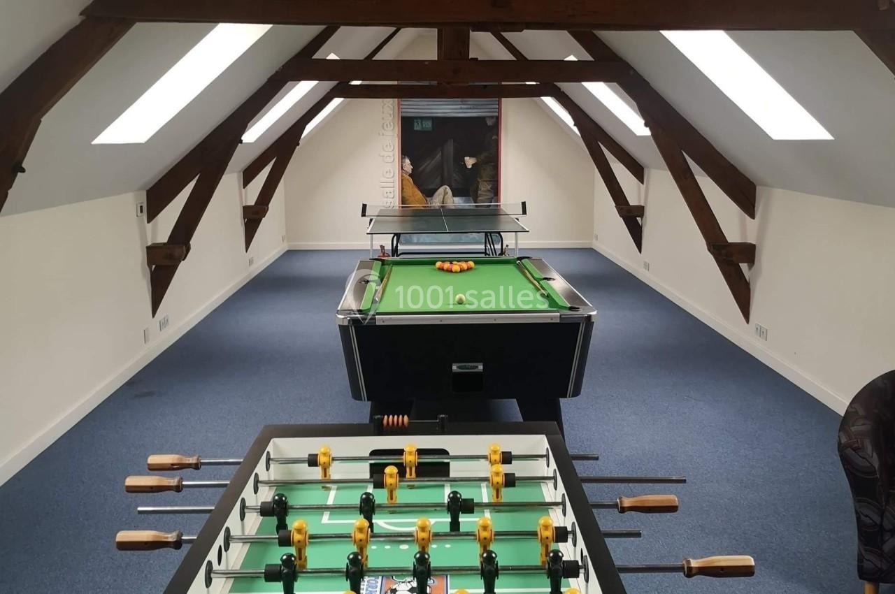 Salle mansardée avec baby-foot et billard alignés sur un sol en moquette bleue, éclairée par des fenêtres de toit.