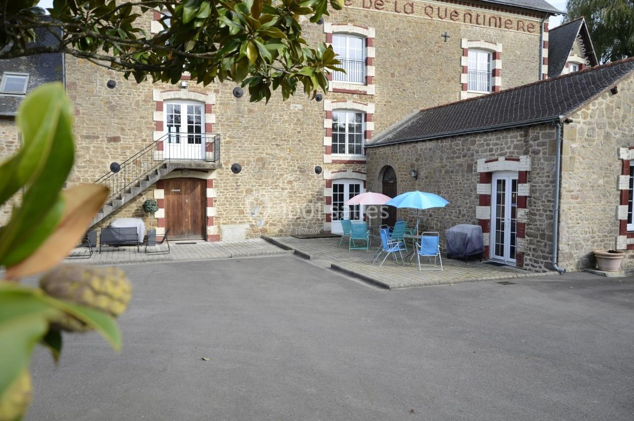 Cour pavée devant une maison en pierre avec des tables, chaises et parasols colorés, entourée de verdure.