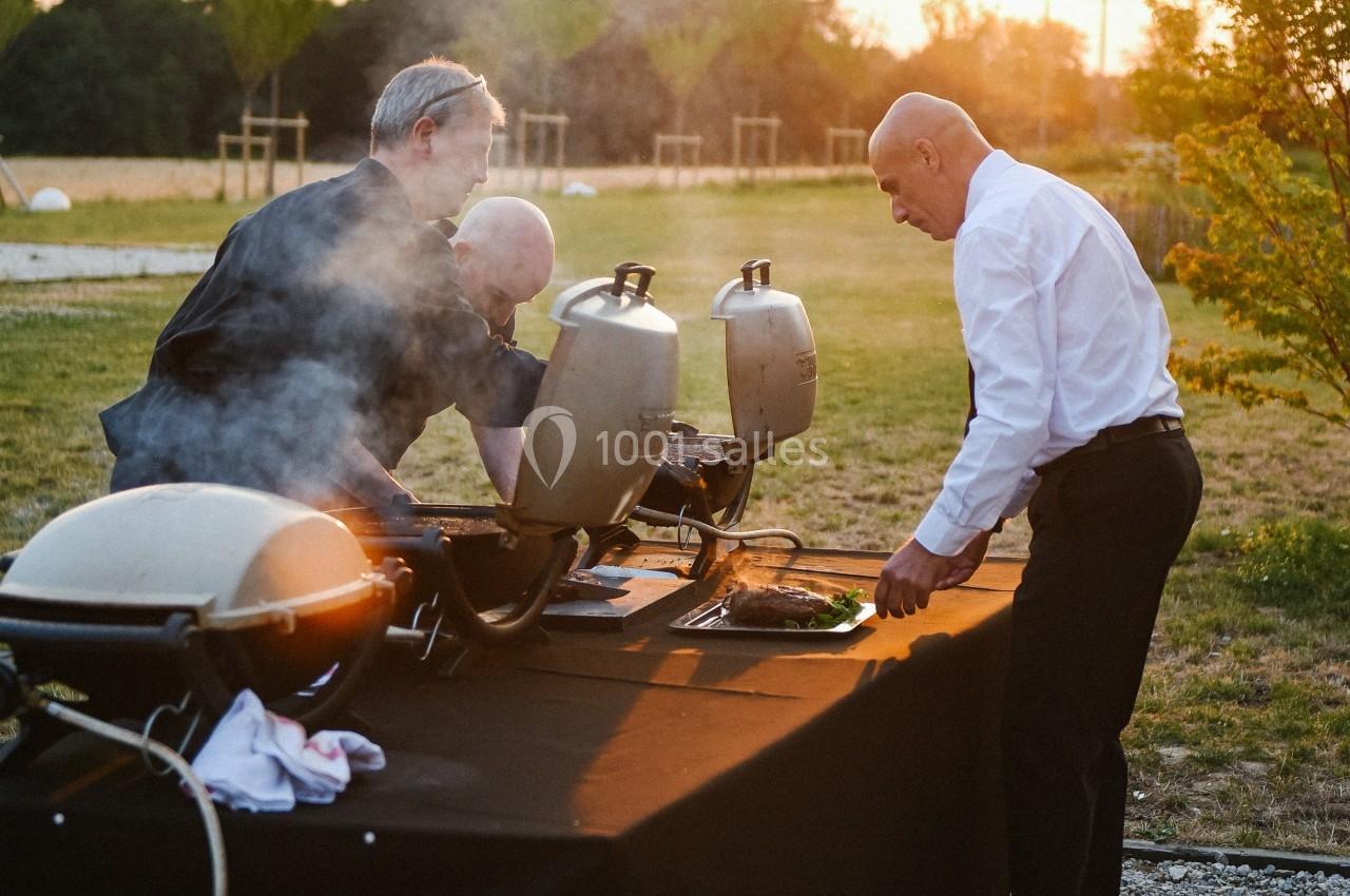Deux hommes cuisinent sur des barbecues en plein air, tandis qu'un troisième surveille la cuisson au coucher du soleil.