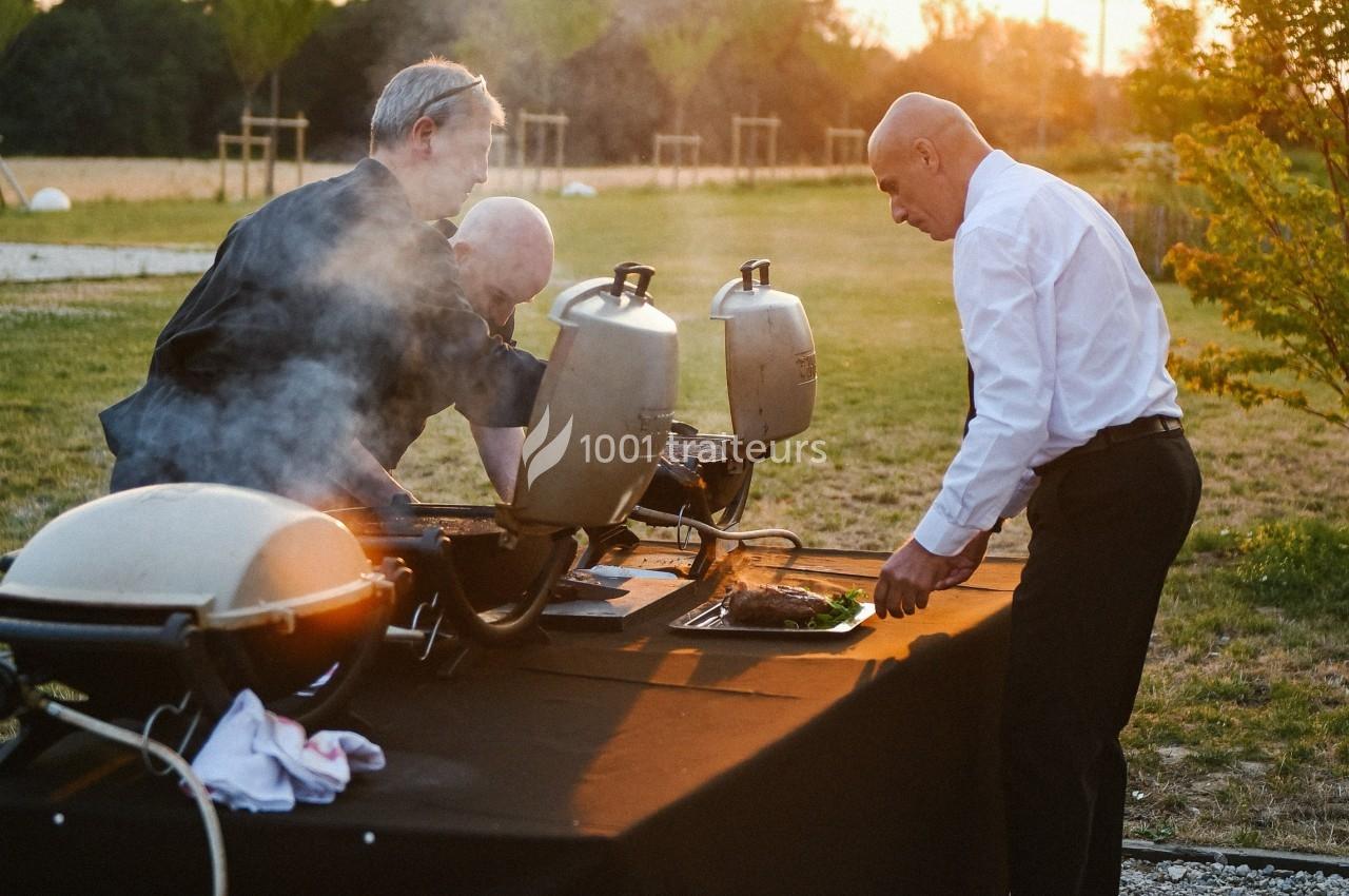 Deux hommes cuisinent au barbecue en plein air, entourés de verdure et éclairés par une lumière de fin de journée.