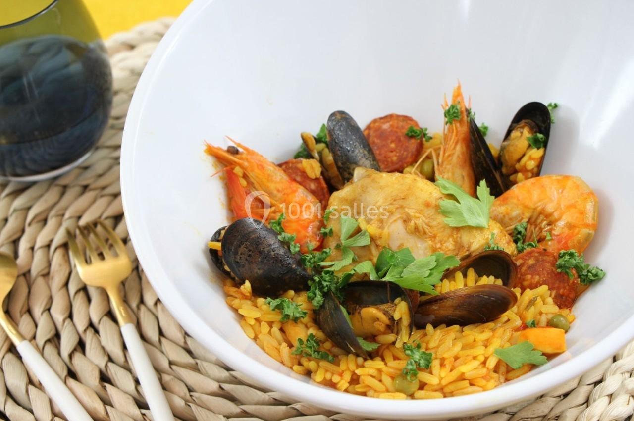 Plat de paella dans un bol blanc, garni de crevettes, moules, poulet, chorizo et persil, posé sur un set tressé.