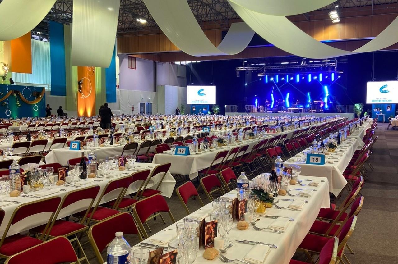 Salle de réception avec de longues tables dressées, des chaises rouges et une scène éclairée en arrière-plan.