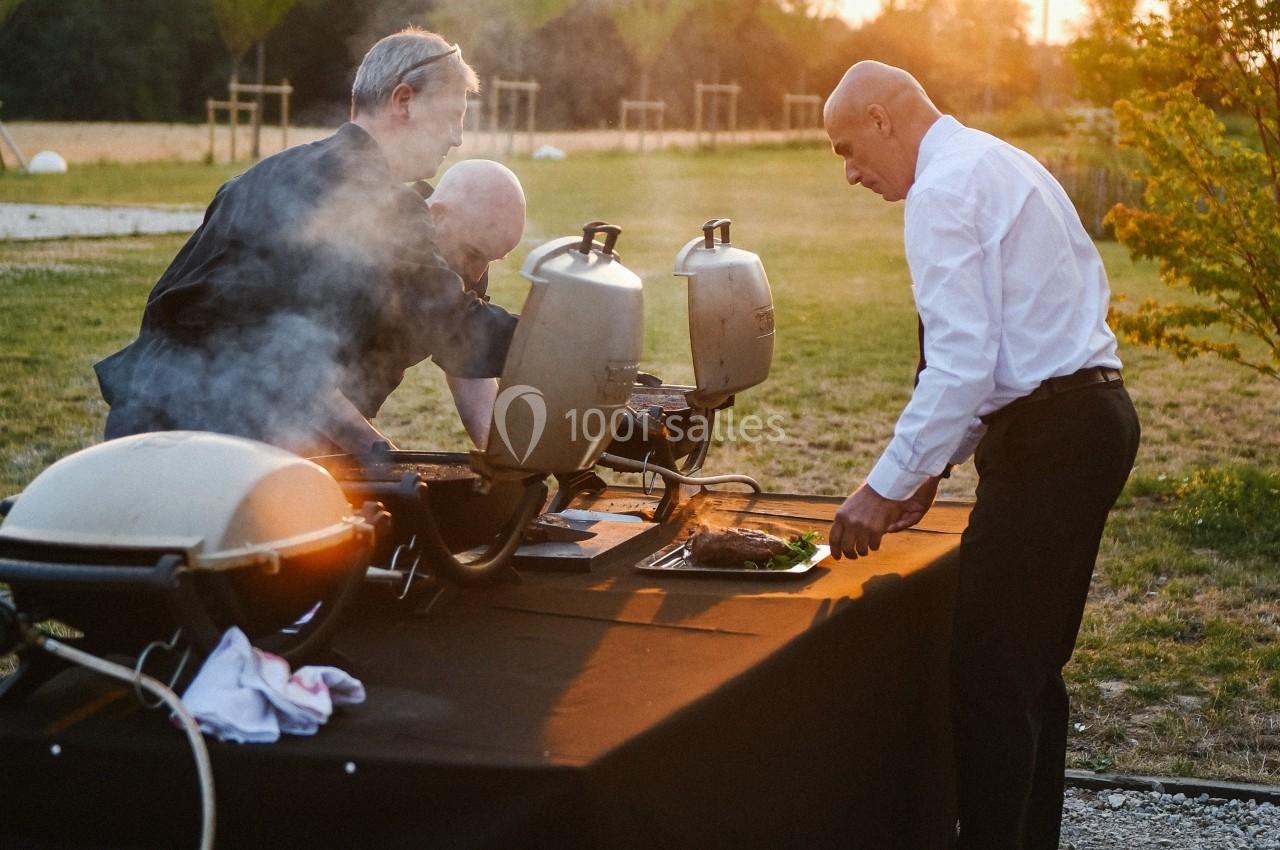 Trois hommes préparent des plats sur des barbecues en plein air, dans un cadre naturel au coucher du soleil.