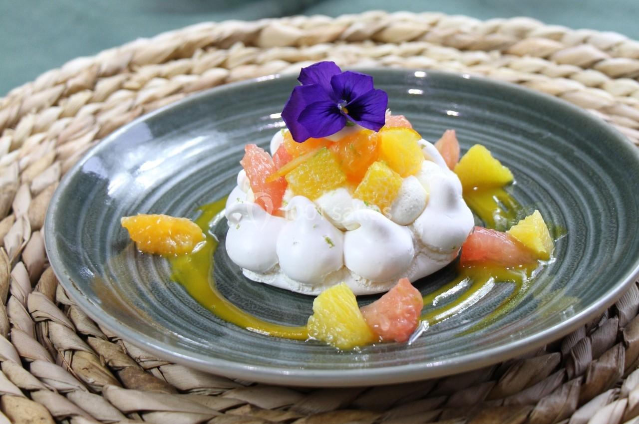 Dessert pavlova garni de morceaux d'agrumes colorés et décoré d'une fleur violette, présenté sur une assiette grise.