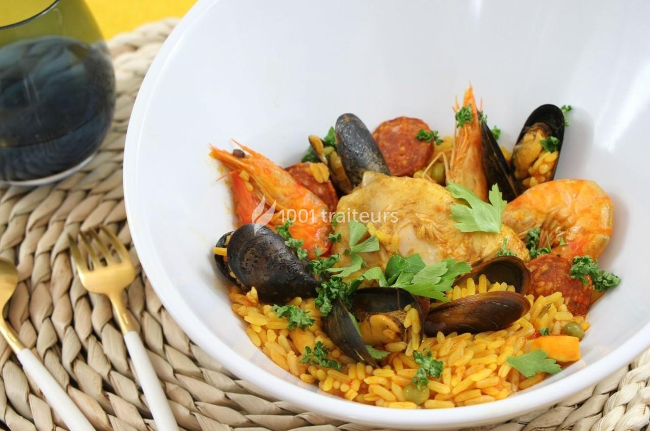 Plat de paella dans un bol blanc, garni de crevettes, moules, poulet, chorizo et persil, posé sur un dessous en osier.