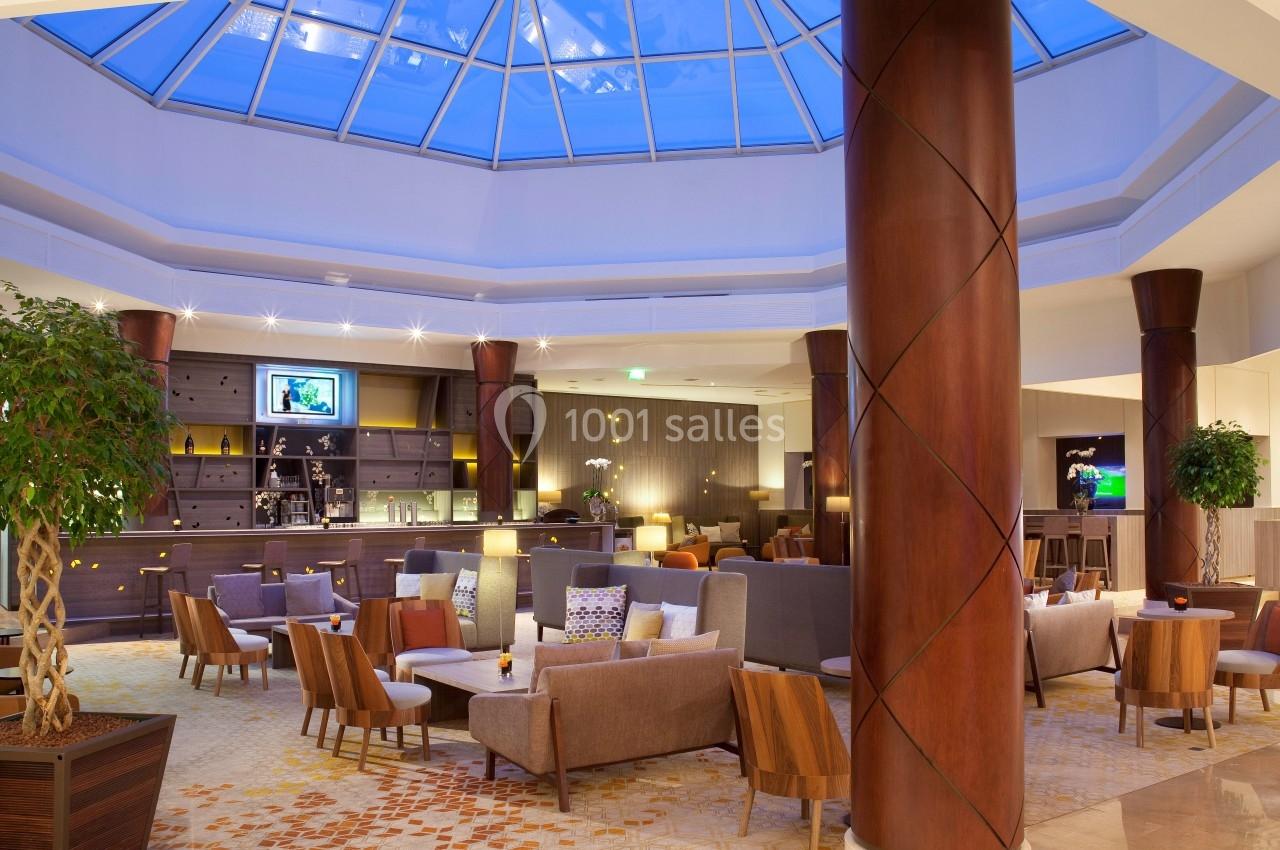 Salon d'hôtel moderne avec fauteuils, tables basses, bar en arrière-plan et verrière laissant entrer la lumière naturelle.